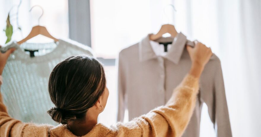 女性が2枚の服を手に取り、どちらを着るか選んでいる様子。明るい室内でのクローゼット整理の場面。