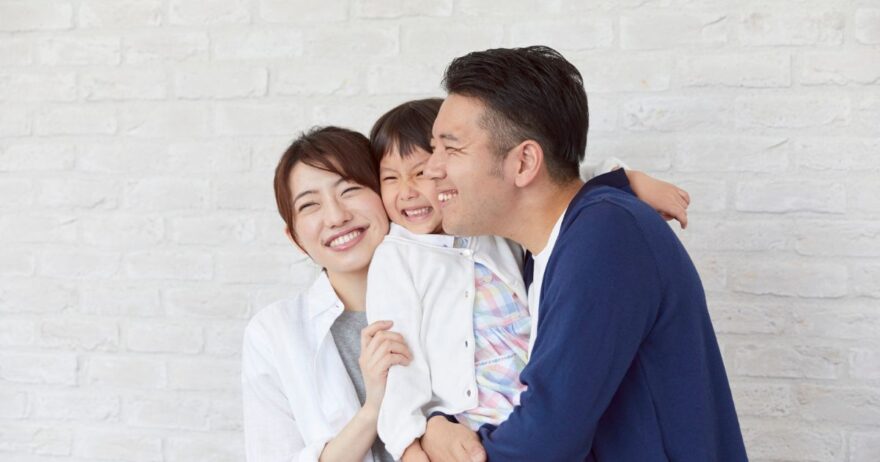 白い壁を背景に、笑顔でハグをする親子3人の家族写真。母親と父親が子供を中央に抱き、笑顔が溢れる家族の幸せな様子が伝わる。