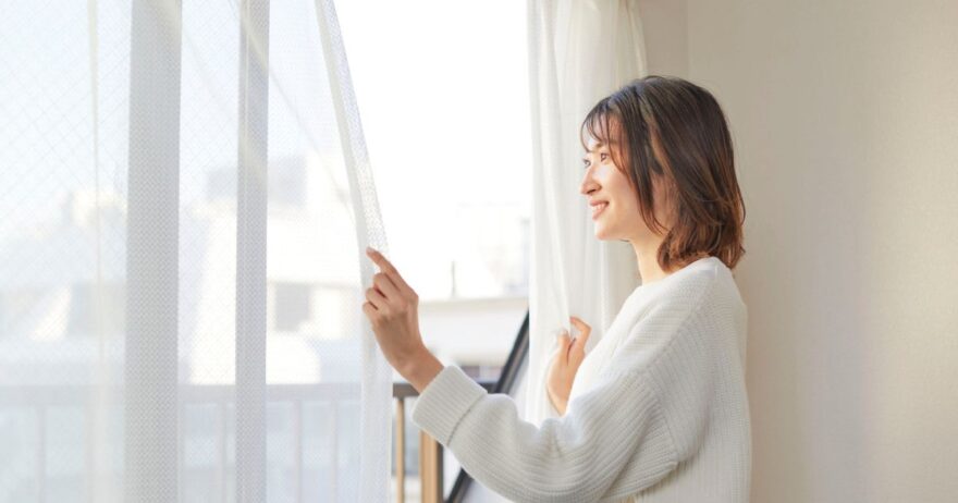 女性が白いカーテンを開けて朝の光を取り入れている様子。白い壁と明るい室内、柔らかな光の中で微笑む女性。