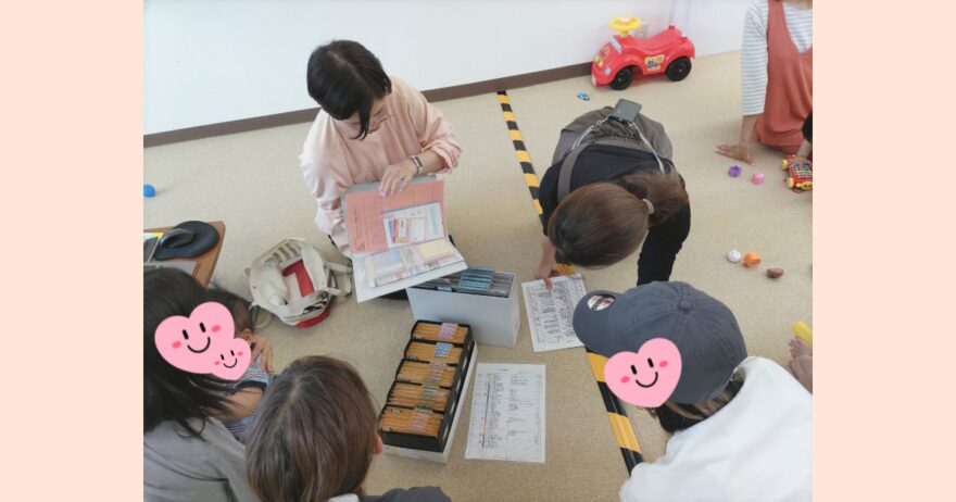 親子のひろばで開催された子育て講座の様子。講師が笑顔で手を振っており、後方では親子が遊びながら話を聞いている。