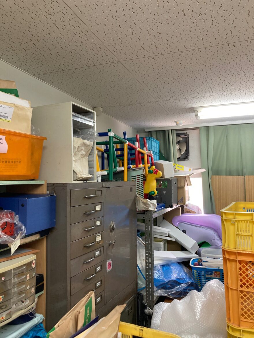 倉庫内にモノが積み上がった状態。棚の上にカラフルな遊具や青いケースが置かれ、雑然とした印象。