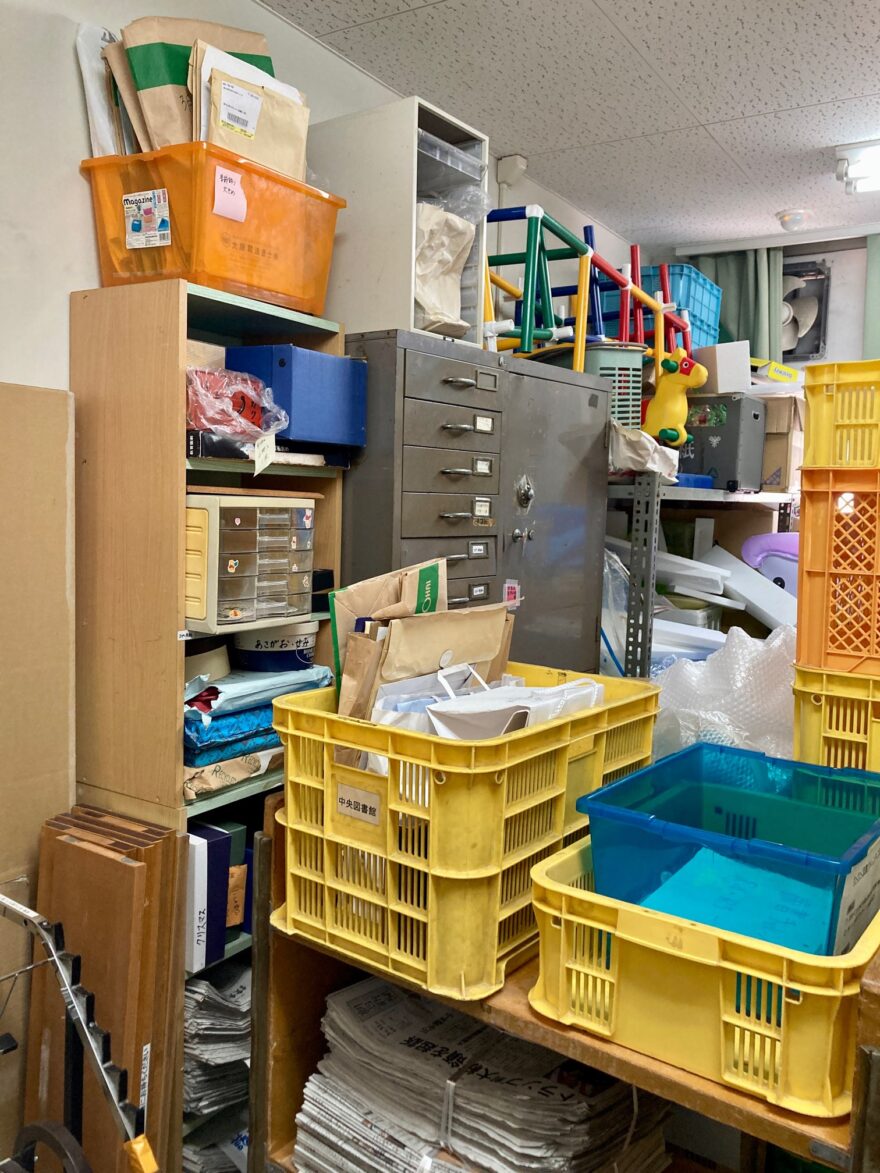 大量の黄色いプラスチックケースや段ボール、新聞紙が積まれた状態の倉庫。通路が狭くなっている様子。
