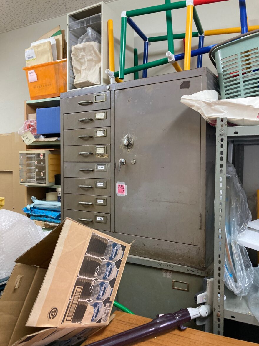 金属製の書類棚の上に遊具や書類ケースが積まれ、全体的にごちゃついた印象の倉庫。
