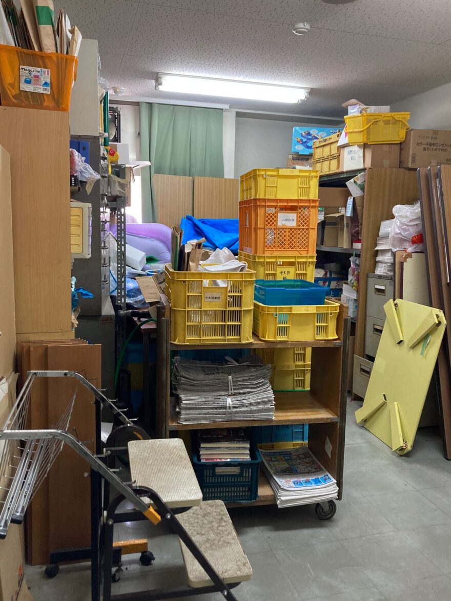 通路に新聞紙やかごが並び、棚の上にもモノがあふれている状態の倉庫。整理前の様子。
