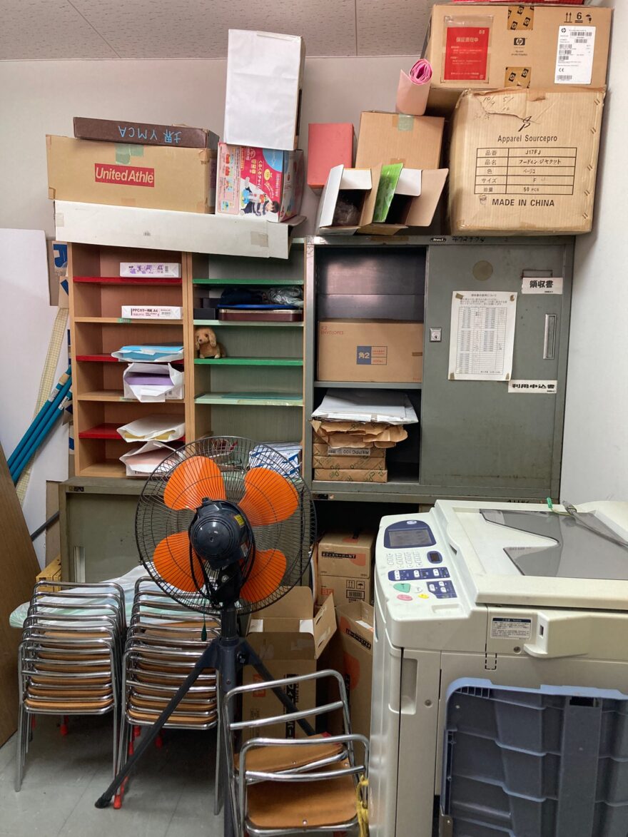 倉庫の一角に積み上げられた段ボールや書類、扇風機、コピー機などが置かれた混在空間。
